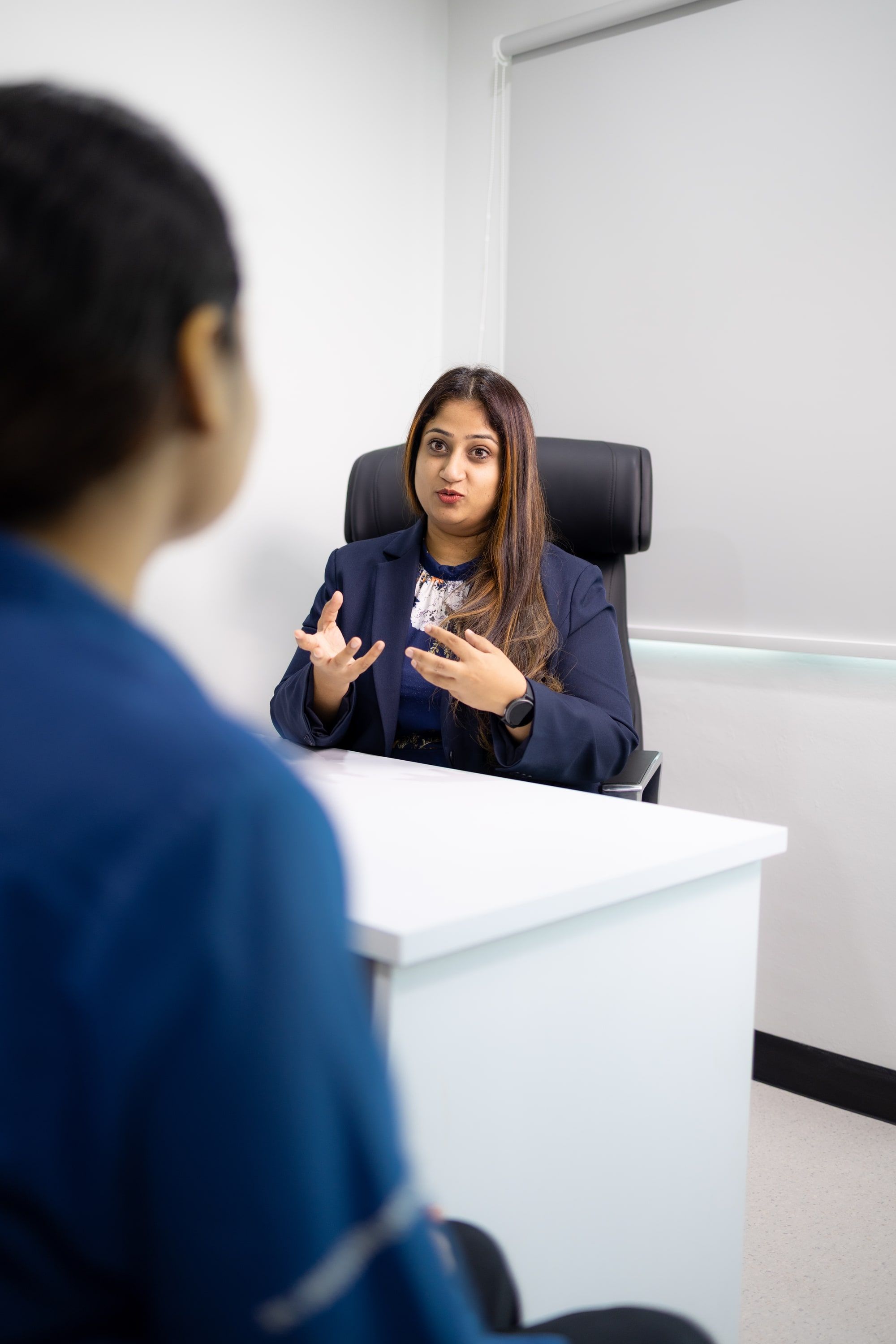 Dr Bheena Vyjhallaja in her consulting room, mid-sentence with both hands raised, talking across the desk.