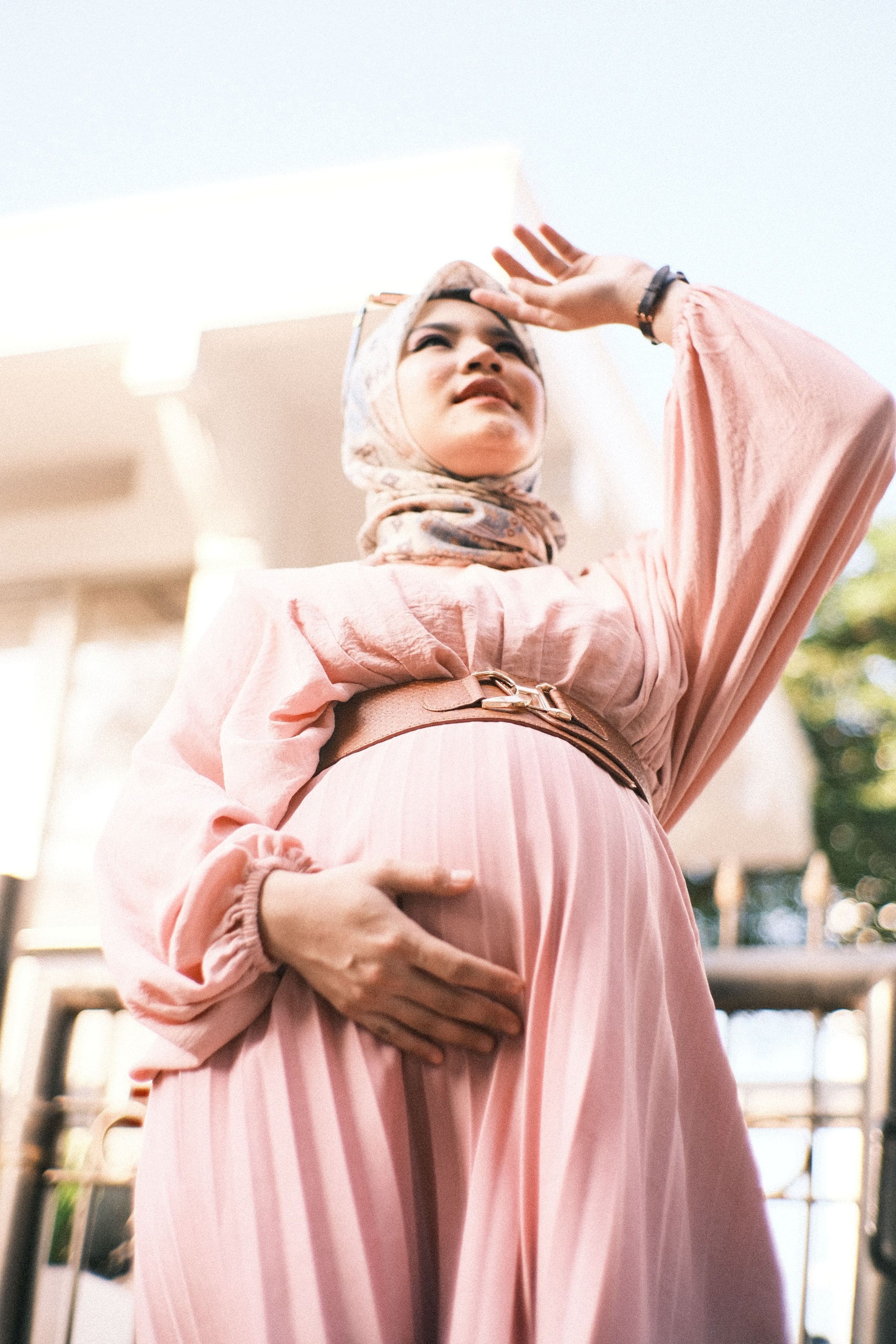 A pregnant woman standing in daylight with both hands resting on her abdomen.